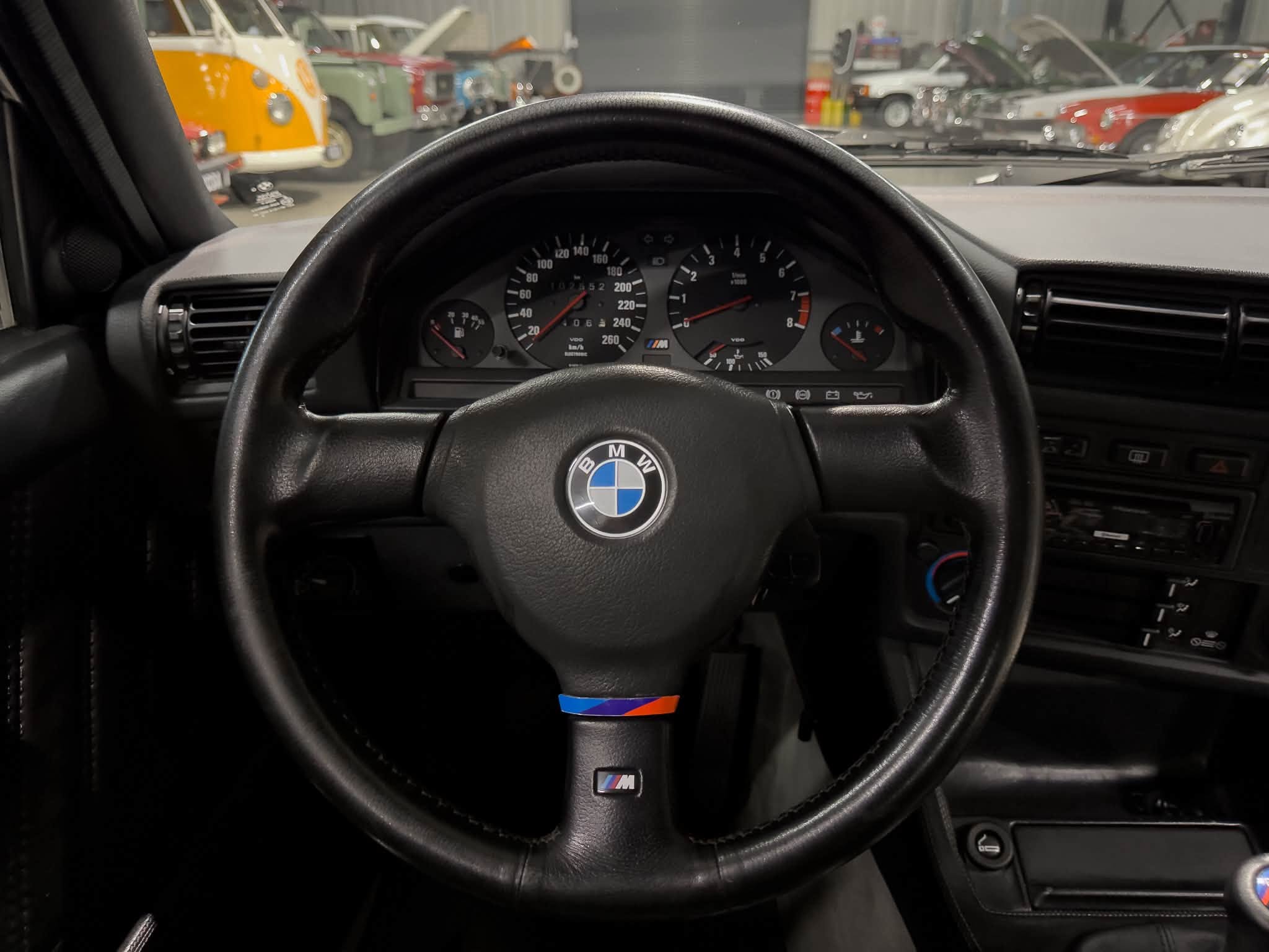 Driver's perspective of a BMW with M badge steering wheel; instrument cluster visible, showing speedometer and tachometer, with a classic car showroom/garage seen through the windshield.